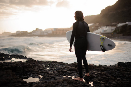 后景黑发男孩站在岩石海滩上，手里拿着冲浪板，看着下的大海。