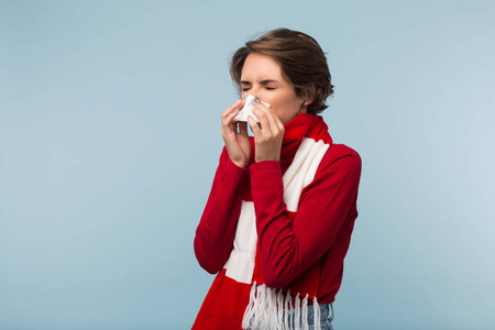 穿着红毛衣,戴着围巾,用餐巾在蓝色背景上吹鼻涕的年轻不健康女性照片