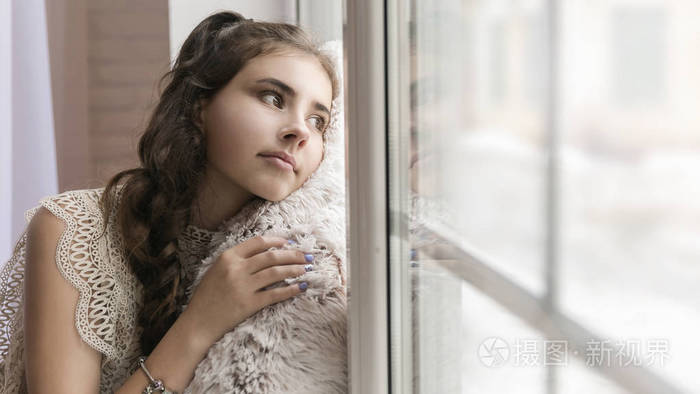 美丽的黑发女孩坐在窗台上,抱着一个柔软的米色枕头,望着窗外.