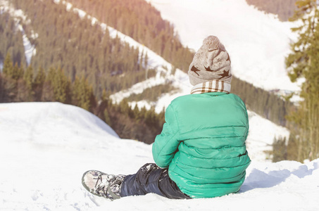 男孩坐在一座覆盖着雪的小山上，背景是松林和山脉。 冬季
