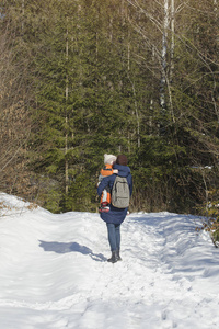 母亲抱着儿子，背包客在针叶林的背景下沿着白雪覆盖的道路行走。 后景。 冬天。 垂直框架