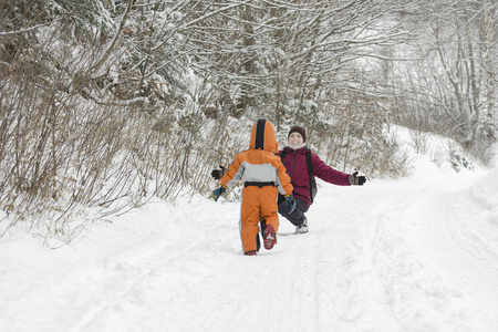 穿着工作服的小男孩穿过雪向母亲跑去。 针叶林的冬日。 背面视图