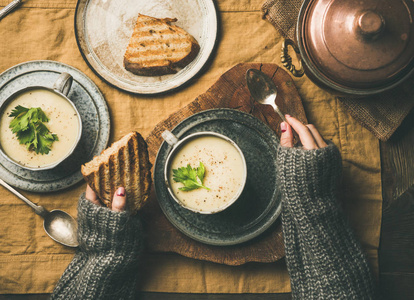 秋天的冬天回家晚餐。 平躺的秋季温暖芹菜奶油汤和女性手与烤面包和勺子在亚麻桌布上的顶部视图。 舒适食品，素食，健康和缓慢的食物概