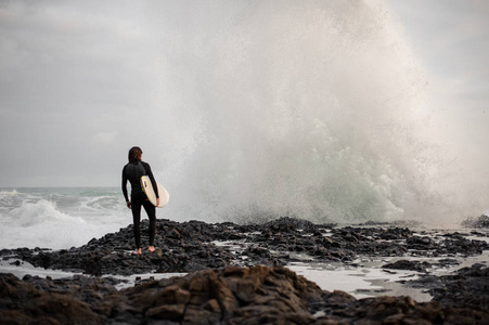 后景黑发男孩站在岩石海滩上，手里拿着冲浪，看着海浪。