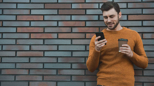 嬉皮士短信短信手机应用程序在城市街道砖墙背景。 惊人的男人拿着智能手机在智能休闲服站立。