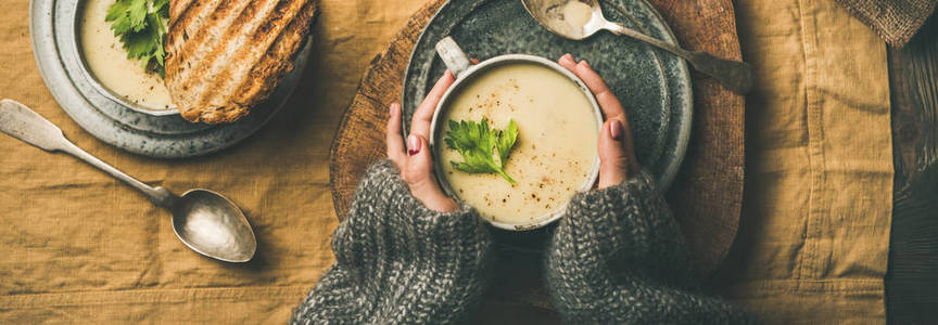 秋天的冬天回家晚餐。 平躺的秋季暖芹菜奶油汤，烤面包和女性手上的亚麻桌布，俯视宽的构图。 舒适食品健康慢食品概念