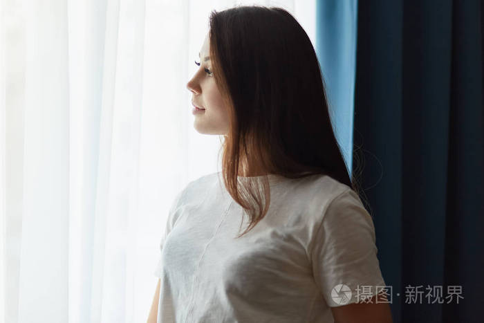 侧身拍摄的体贴的黑发女人穿着白色t恤思考什么,而站在窗边享受平静的