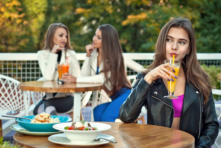 女朋友锻炼后在咖啡馆吃饭。 桌子上有新鲜的果汁和沙拉。 概念健康的生活方式。 穿运动服的女孩。 舒适的户外餐厅和绿色背景。