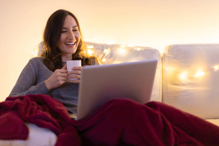 年轻微笑的女人坐在沙发上，拿着笔记本电脑和一杯咖啡或茶，看着屏幕上的放松与和平的概念