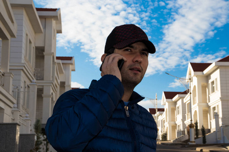 一名房地产经纪人在住宅别墅前，通过手机与潜在客户交谈，背景是一个蓝色多云的天空，中午。