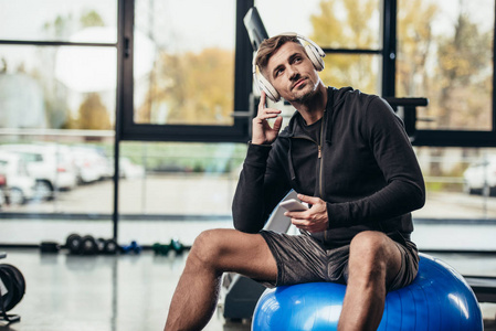英俊的运动员坐在健身球上，在健身房里用智能手机听音乐