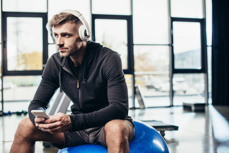 帅哥运动员在健身房里坐着带智能手机的健身球