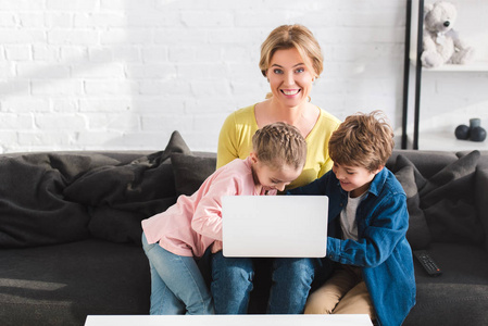 妈妈开心地在相机前微笑，在家里和孩子一起使用笔记本电脑