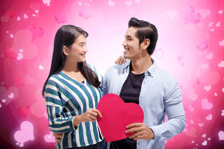 快乐的亚洲夫妇拥抱并抱着红色的纸心在粉红色的背景上。 爱情观念和情人节
