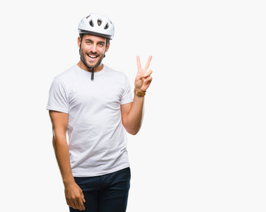 年轻的帅哥戴着自行车安全头盔，在孤立的背景上微笑，快乐的脸对着镜头眨着眼睛，做着胜利的标志。 二号。