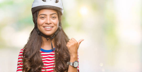 年轻的阿拉伯骑自行车的女人戴着安全帽，在孤立的背景上微笑，快乐的脸看着，用拇指指向一边。