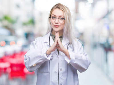 年轻的金发医生女人在孤立的背景下祈祷，双手一起请求宽恕，微笑自信。