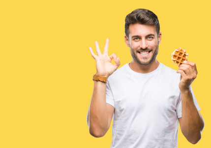 年轻的帅哥，在孤立的背景上吃甜华夫饼，用手指做OK标志，这是极好的象征。
