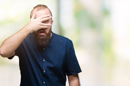 年轻的白种人臀部男子在孤立的背景下偷看震惊，覆盖着脸和眼睛，用手透过手指看着尴尬的表情。