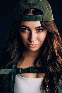 军事时尚。 特写美丽黑发女人的肖像，看着黑色背景上的相机。 戴着深绿色帽子的漂亮女孩看着摄像机