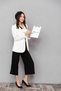 一位穿着夹克和裤子的微笑的年轻女子站在灰色背景上的全长画像，展示了一张带有图表的平板电脑