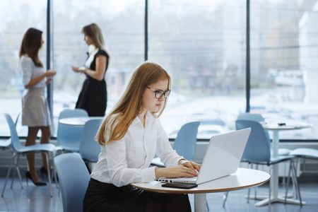 办公室工作人员团队。 一个长头发的女孩戴着眼镜和一件白色衬衫坐在一张桌子上，桌上有一台笔记本电脑，两个商务女性站在后台说话。 团