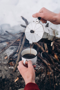 旅游男子A坐在营地附近的火安在冬天，并倒自己热咖啡。 概念冒险活动户外度假。 冬季野营