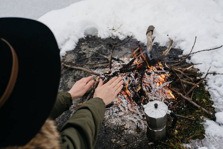 戴帽子的女孩在冬天的火灾附近温暖双手。 概念冒险活动户外度假。 冬季野营