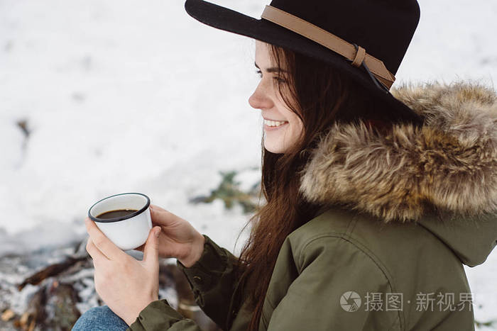 在冬天，戴着帽子的快乐女孩坐在篝火旁，喝着一杯热咖啡。 概念冒险活动户外度假。 冬季野营