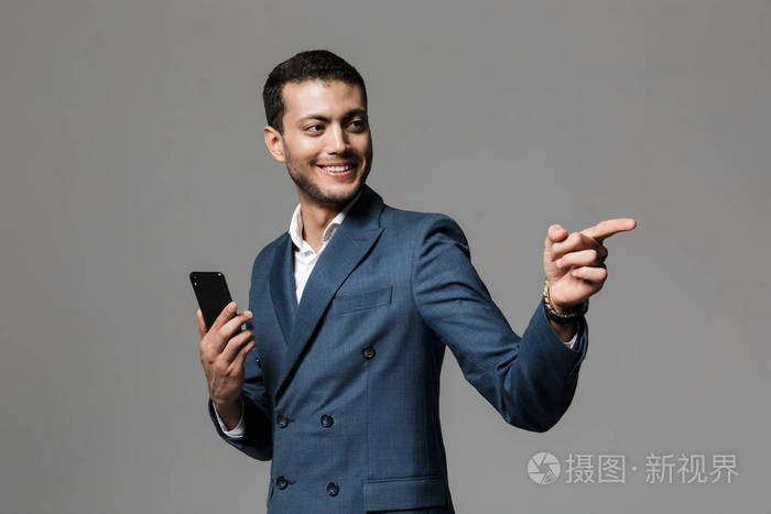 一位微笑的年轻商人的肖像，他穿着西装，站在灰色背景下，手里拿着手机，指着远处