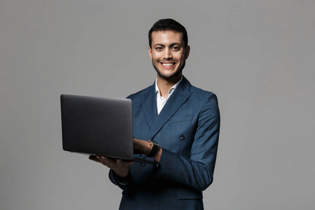 一位微笑的年轻商人的肖像，他穿着西装，用笔记本电脑在灰色背景下孤立地站立着