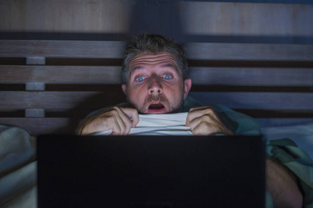 生活方式肖像年轻的恐惧和压力男人躺在床上，深夜用笔记本电脑或卧室电视观看互联网恐怖电影，惊恐的表情覆盖着毯子