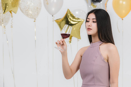 迷人的年轻亚洲女人穿着粉红色的连衣裙，拿着她的饮料，高兴地在白色背景上的有趣的派对肖像与五颜六色的节日气球。