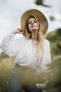 穿着草帽白色亚麻上衣和牛仔裤的时髦女人，摆在乡间草地上