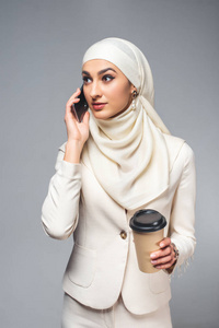 年轻的穆斯林妇女用智能手机说话，拿着灰色的纸杯