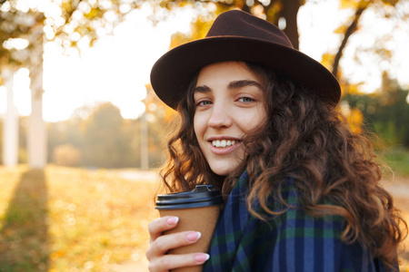 美丽的年轻女子戴着帽子在公园散步，秋天拿着一杯外卖咖啡