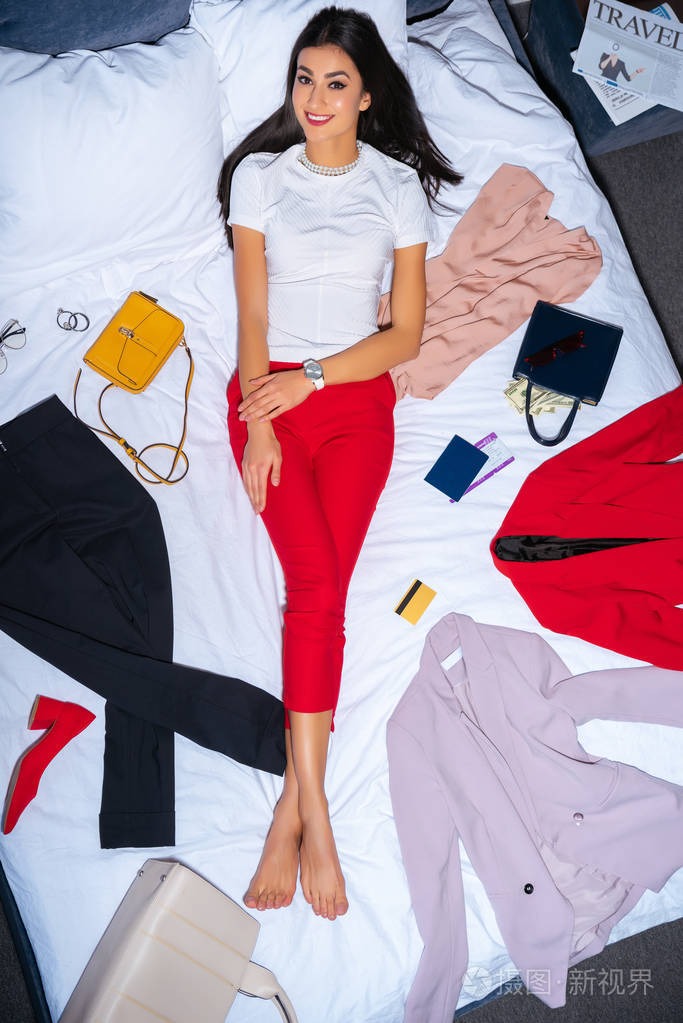 美女躺在床上穿着时髦的衣服护照信用卡和旅行报纸对着镜头微笑