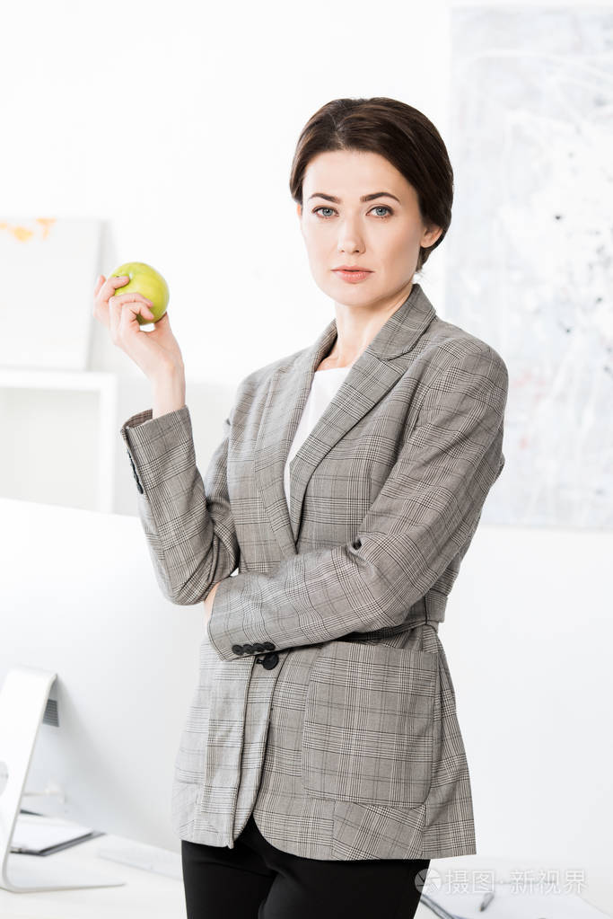 漂亮的女商人穿着灰色西装拿着苹果，在室里看着摄像机