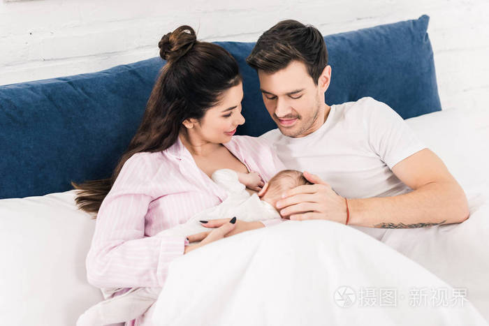 微笑的母亲的肖像,在家中与丈夫一起在床边哺乳小婴儿