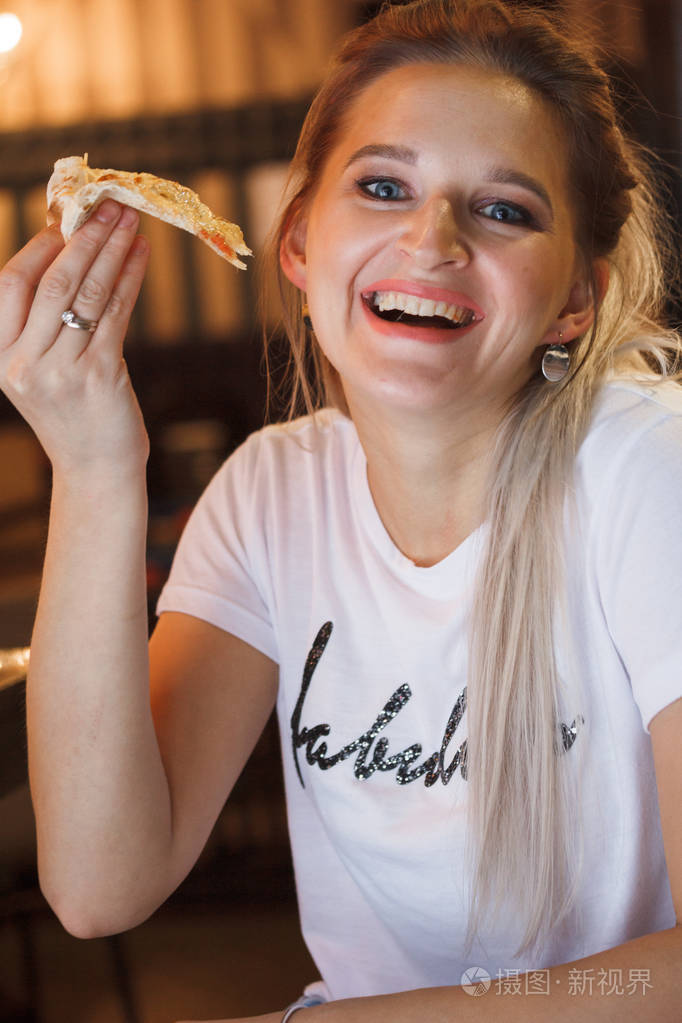 休闲女孩在餐厅吃披萨享受这个穿白色t恤的金发女郎在餐厅吃披萨