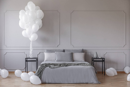 在宽敞的卧室里放白色气球，配有带灰色羽绒被的特大床和黑色床头柜之间的枕头，在空灰墙上有复印空间的真实照片