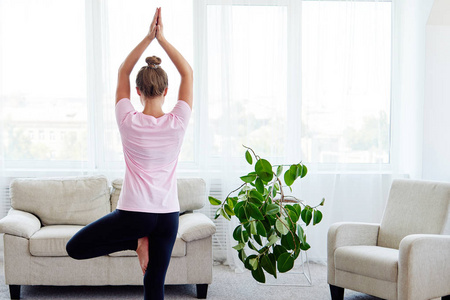 年轻女性练习平衡瑜伽的肖像，在家室内复制空间后景。 做树姿的女孩。 放松和做瑜伽。 健康和健康的生活方式