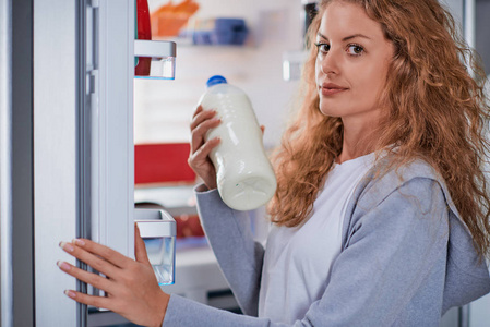 女人站在冰箱前吃牛奶。 装满杂货的冰箱。