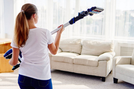 年轻女子的肖像，穿着白色衬衫和牛仔裤，手里拿着无线吸尘器，在家里的客厅里打扫。 家务清洁和家务观念