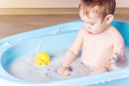 可爱的男婴在浴室里洗蓝色的澡。 这个孩子正在玩黄色的鸭子和肥皂泡沫。