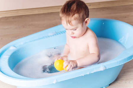 可爱的男婴在浴室里洗蓝色的澡。 这个孩子正在玩黄色的鸭子和肥皂泡沫。