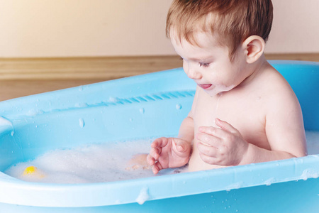 可爱的婴儿在浴室的蓝色浴缸里洗澡。 男孩在玩水和肥皂泡沫