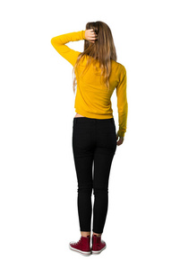 一个年轻女孩穿着黄色毛衣的背部姿势向后看，同时在孤立的白色背景上挠头