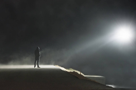 一个人站在一条河边一个孤独的戴着兜帽的人站在一条朦胧的路上,看着