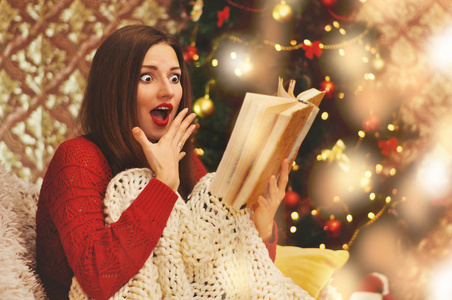 一个穿着红色毛衣的漂亮受惊的女人坐在沙发上的针织毯子下面，读着一本旧书。 背景中花环上的圣诞树。
